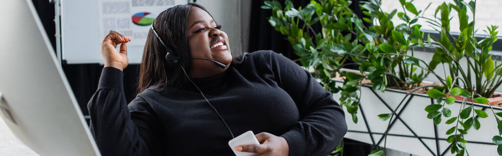 Mutlu Afro-Amerikan artı boyut operatörü mikrofonla konuşuyor ve akıllı telefonu ofisinde tutuyor. - Fotoğraf, Görsel