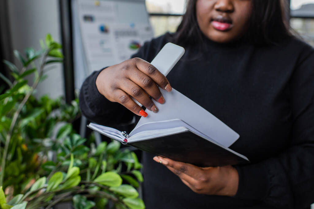 vista ritagliata di africano americano plus size donna in possesso di notebook e smartphone in ufficio - Foto, immagini