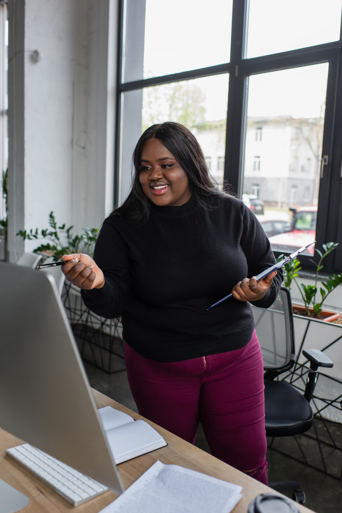 uśmiechnięty african amerykański plus rozmiar businesswoman trzyma schowek i długopis w pobliżu monitora komputerowego w biurze - Zdjęcie, obraz