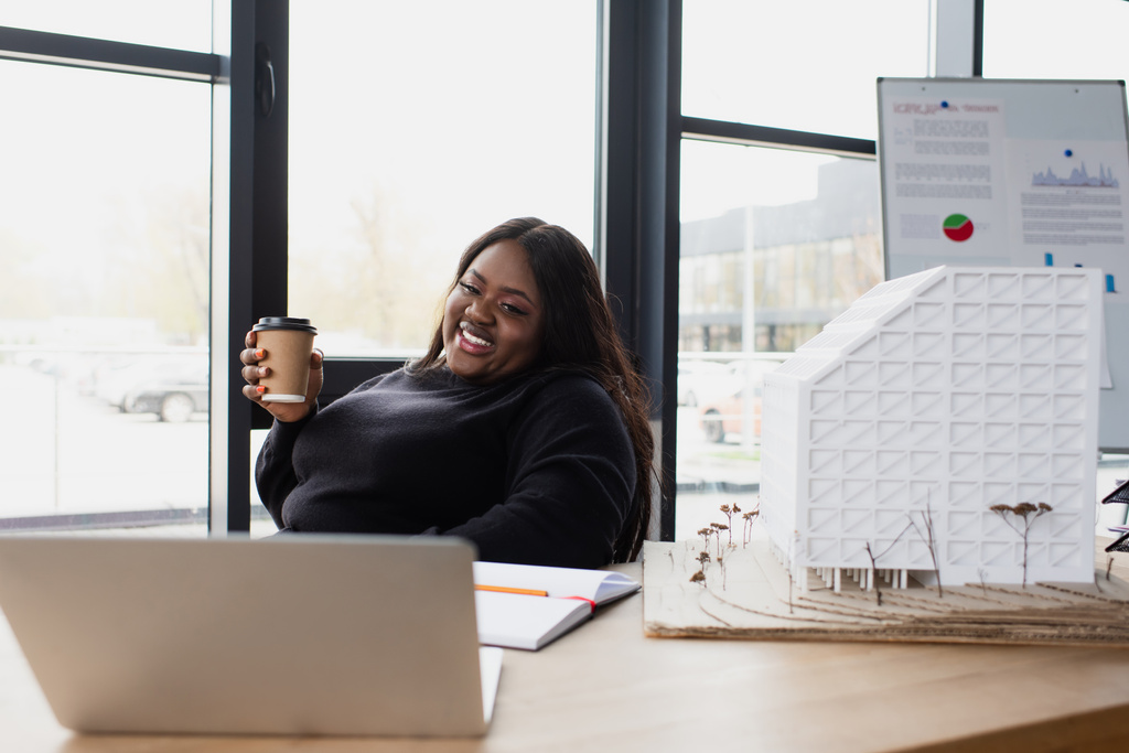 heureux afro-américain plus taille ingénieur tenant tasse en papier tout en regardant ordinateur portable près modèle de maison  - Photo, image