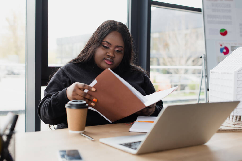 Afrikanisch-amerikanische Plus-Size-Frau blickt auf Ordner in der Nähe von Gadgets und Pappbecher auf dem Schreibtisch - Foto, Bild