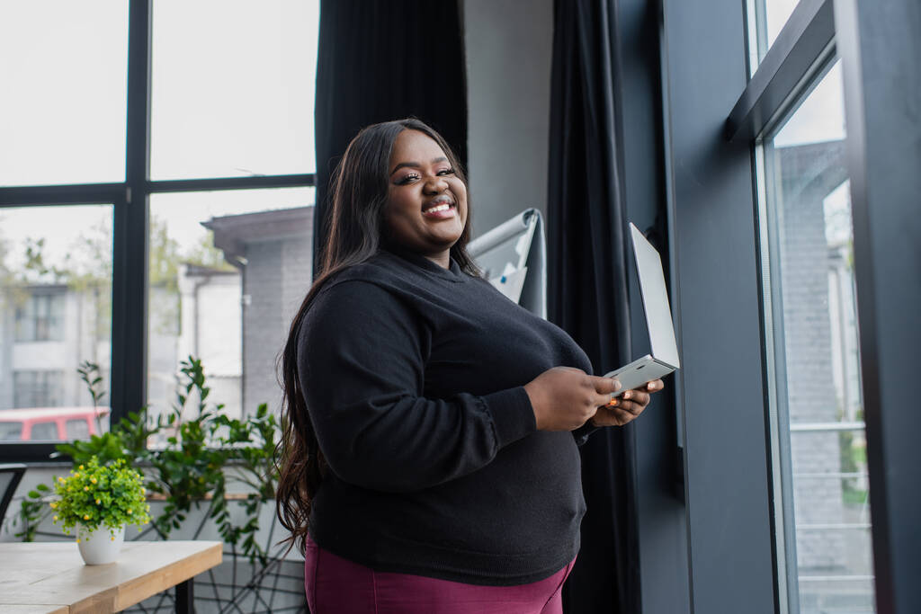 χαρούμενη Αφροαμερικανίδα συν μέγεθος επιχειρηματίας κρατώντας φορητό υπολογιστή κοντά στο παράθυρο στο γραφείο  - Φωτογραφία, εικόνα