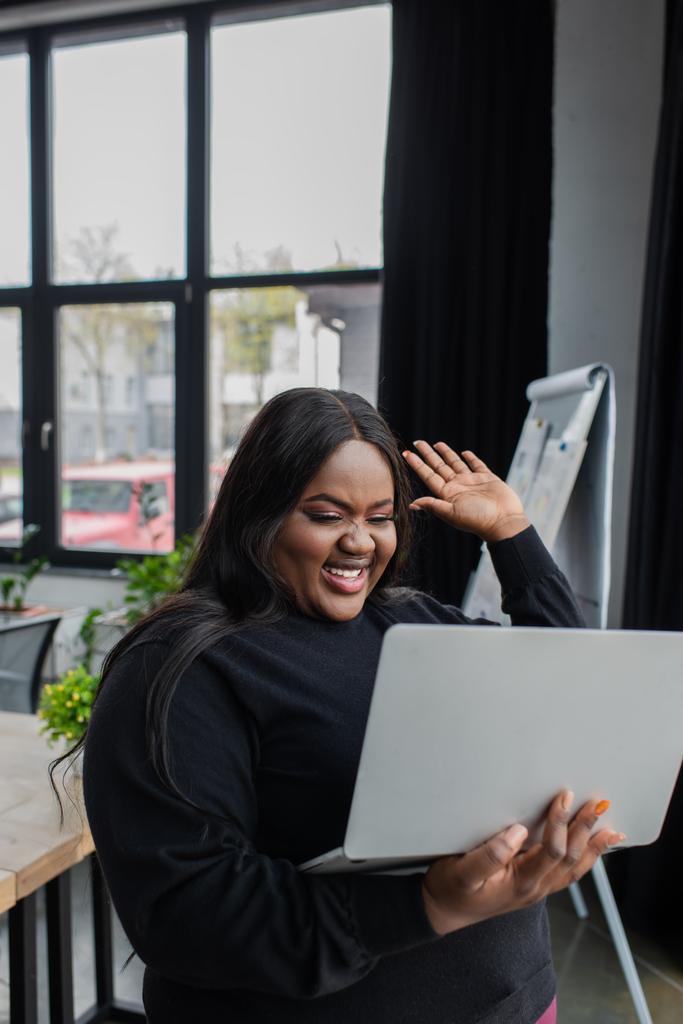 χαρούμενη Αφροαμερικάνα συν μέγεθος επιχειρηματίας κρατώντας φορητό υπολογιστή και χαιρετώντας το χέρι, ενώ έχοντας βιντεοκλήση  - Φωτογραφία, εικόνα