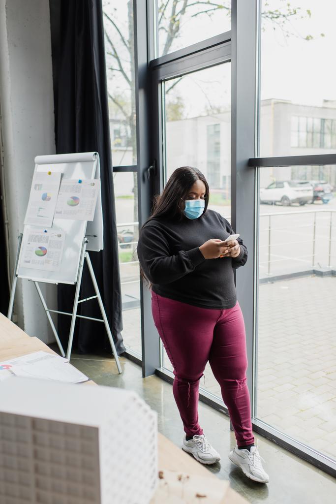 afro-américaine plus la taille femme d'affaires dans le masque médical en utilisant un smartphone près du modèle de maison floue  - Photo, image