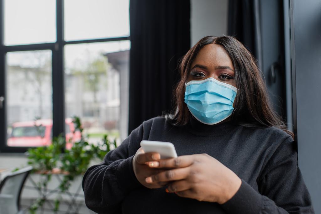 Αφροαμερικάνα συν μέγεθος επιχειρηματίας σε ιατρική μάσκα χρησιμοποιώντας smartphone στο γραφείο - Φωτογραφία, εικόνα