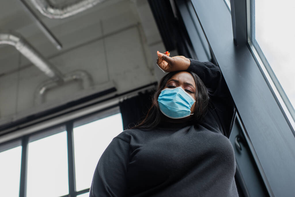 Niedrigwinkel-Ansicht einer afrikanisch-amerikanischen Plus-Size-Unternehmerin mit medizinischer Maske, die sich im Büro unwohl fühlt - Foto, Bild