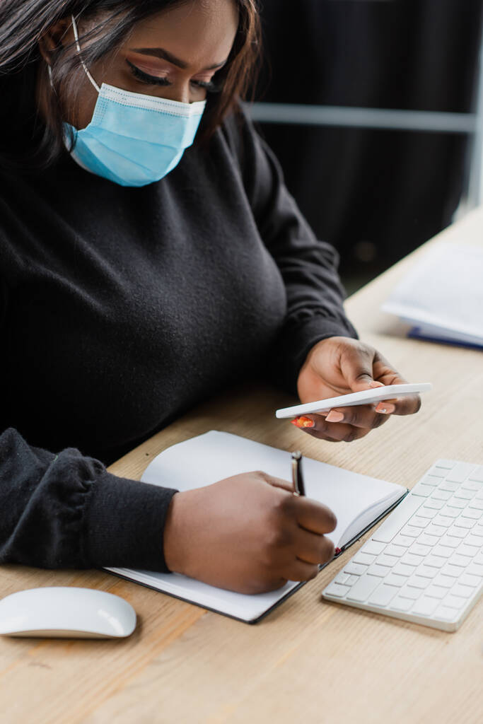 Африканский американец плюс размер деловая женщина в медицинской маске держа смартфон во время записи в ноутбуке  - Фото, изображение