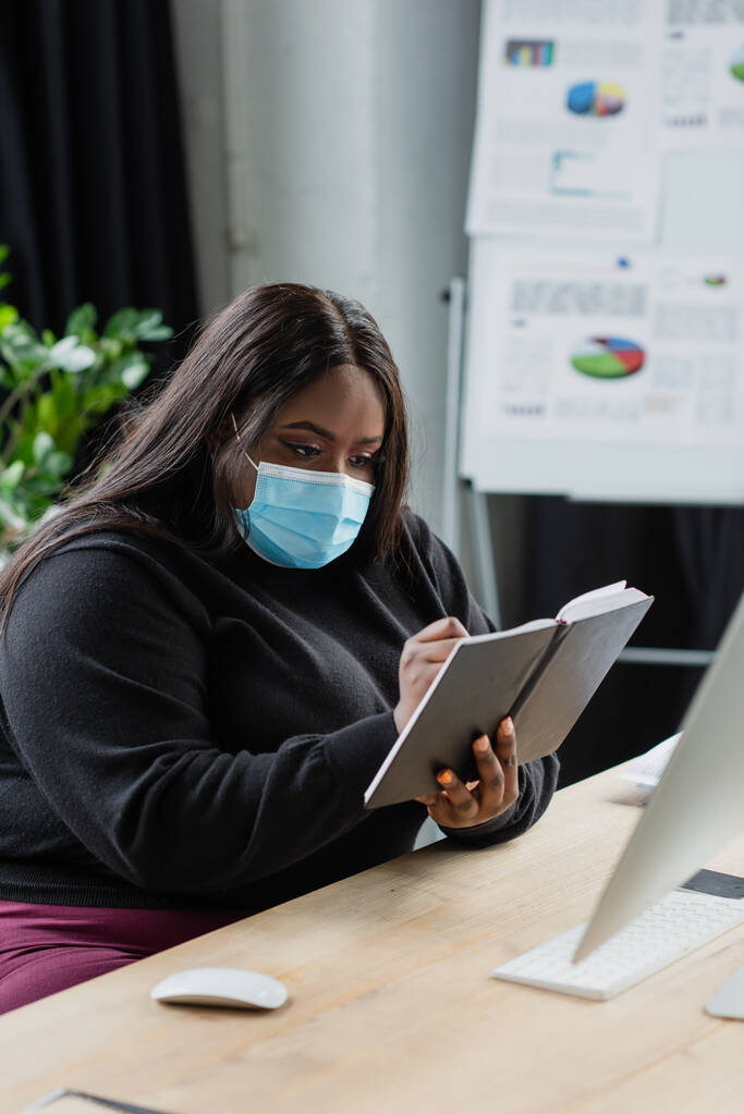 afroamericano plus size donna d'affari in maschera medica in possesso di notebook vicino al computer  - Foto, immagini
