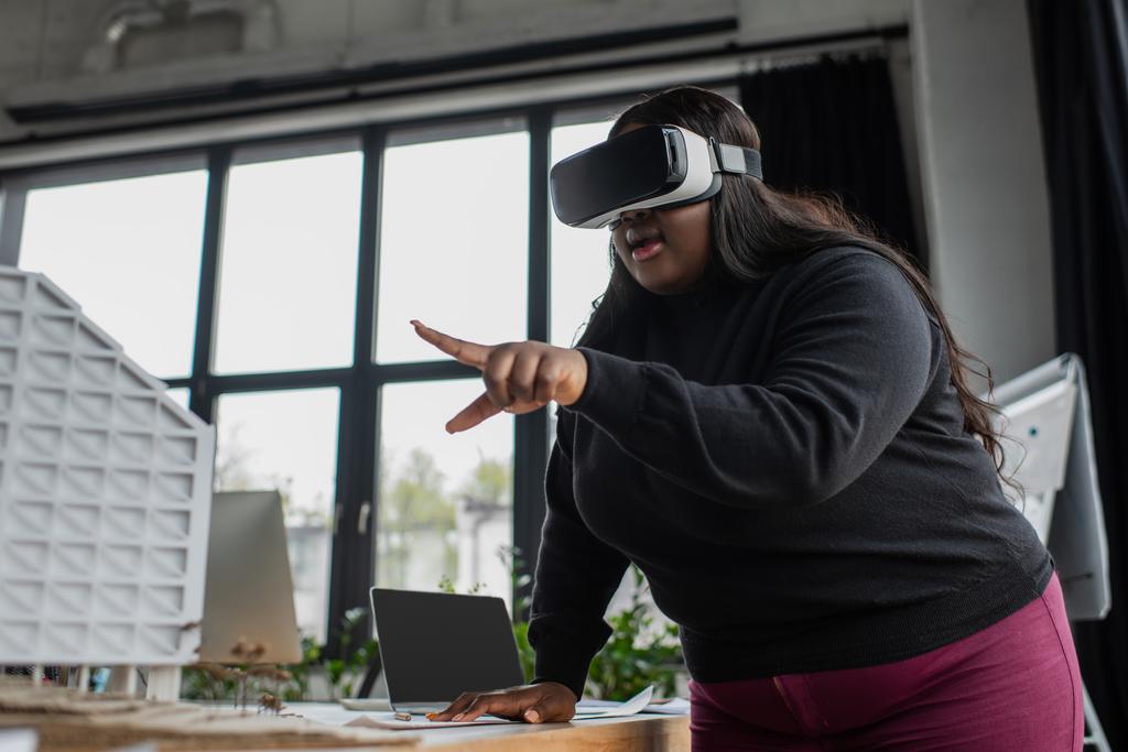 έκπληκτος αφρικανός αμερικανός συν μέγεθος μηχανικός σε VR ακουστικά που δείχνουν στο μοντέλο χάρτινο σπίτι  - Φωτογραφία, εικόνα