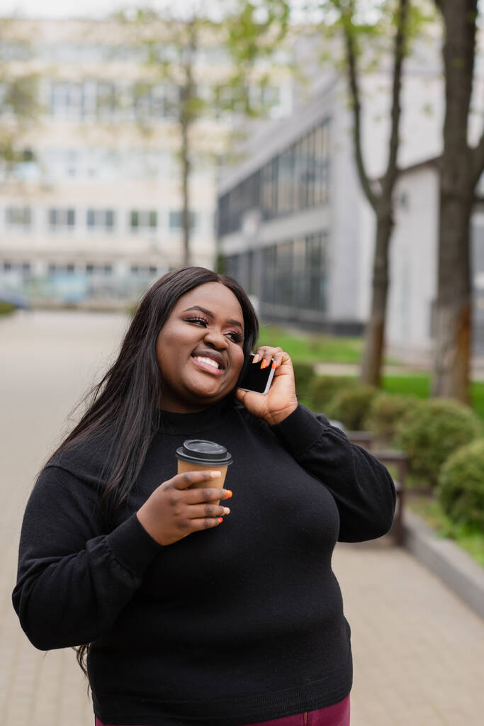 весела афроамериканка плюс розмір жінка тримає каву, щоб піти і розмовляти по смартфонах зовні
  - Фото, зображення