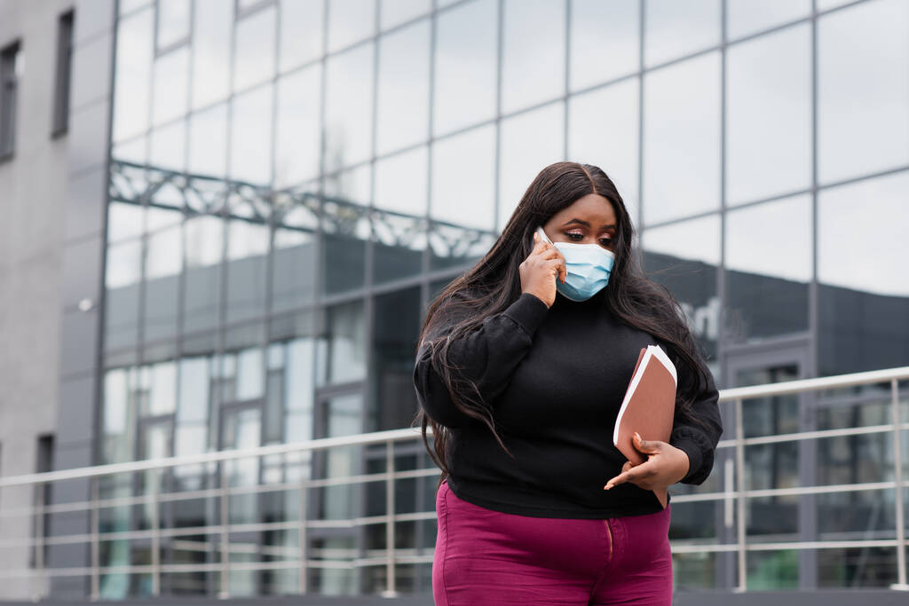 Afrikaans amerikaans plus size zakenvrouw in medisch masker holding map en praten op smartphone buiten  - Foto, afbeelding