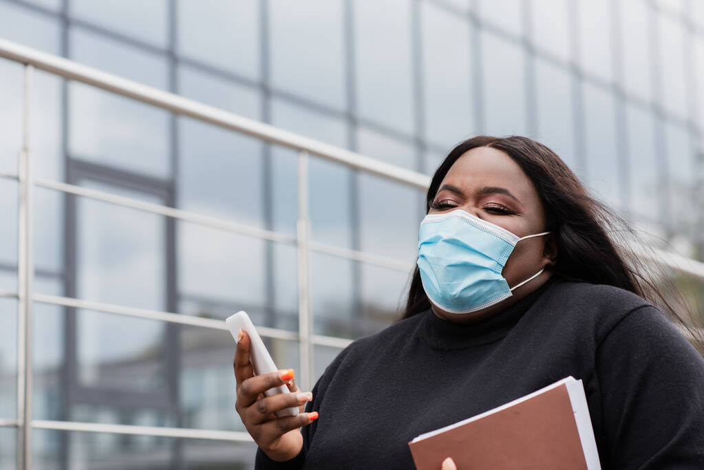 афроамериканська жінка плюс розмір жінка в медичній масці тримає папку і мобільний телефон зовні
  - Фото, зображення
