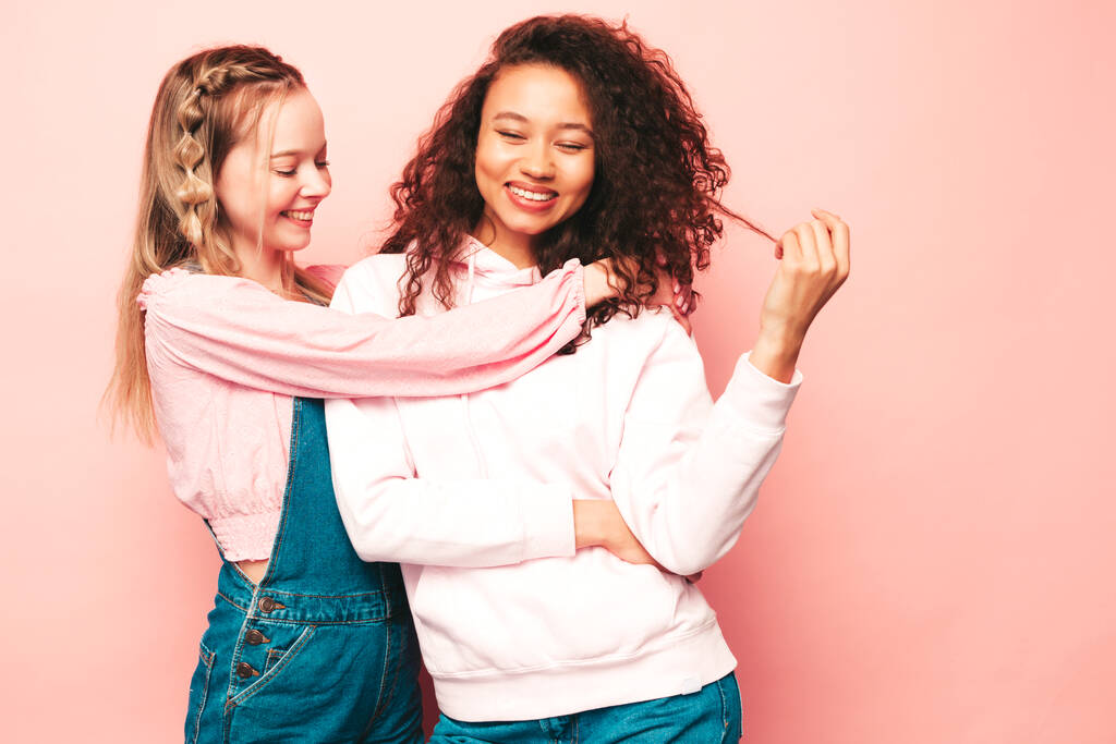 Zwei junge, schön lächelnde internationale Hipsterinnen in trendiger Sommerkleidung. Sexy unbeschwerte Frauen posieren in der Nähe einer rosafarbenen Wand im Studio. Positive Modelle, die Spaß haben. Konzept der Freundschaft - Foto, Bild