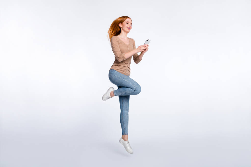 Full length φωτογραφία του αστείο γοητευτικό τζίντζερ κυρία φορούν μπεζ πουκάμισο χαμογελώντας άλμα ψηλά κρατώντας σύγχρονη συσκευή απομονωμένο λευκό φόντο χρώμα - Φωτογραφία, εικόνα