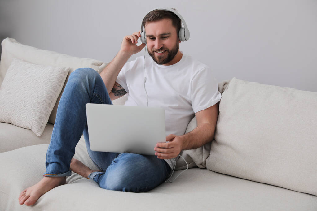 Uomo con laptop e cuffie seduto sul divano di casa - Foto, immagini