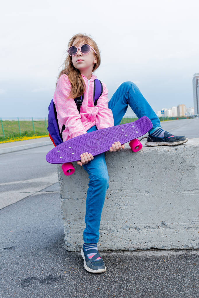 Chica joven en jeans y suéter rosa sostiene un monopatín en la calle. Concepto de estilo de vida adolescente. Copiar espacio. Mochila púrpura en la parte posterior. - Foto, Imagen
