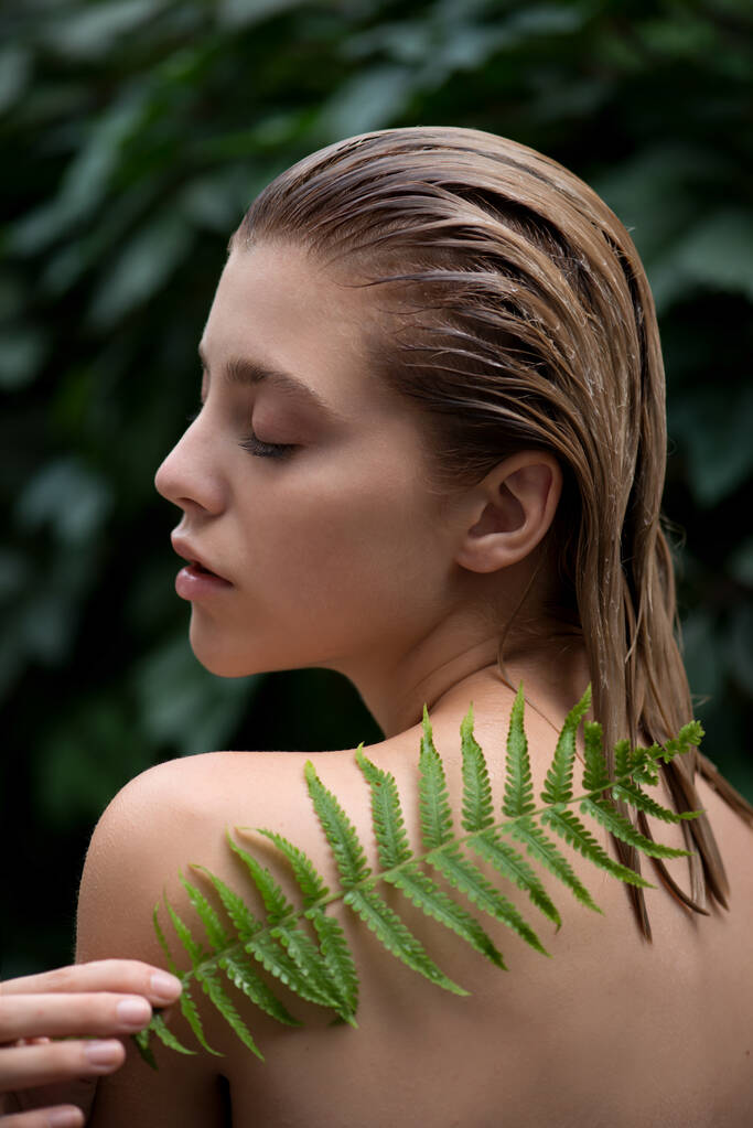 Mooie jonge vrouw met perfecte huid voorkant van plant tropische groene bladeren achtergrond met varen. Model brengt een balsem, shampoo of conditioner aan op haar haar SPA, wellness, lichaamsverzorging en huidverzorging. - Foto, afbeelding