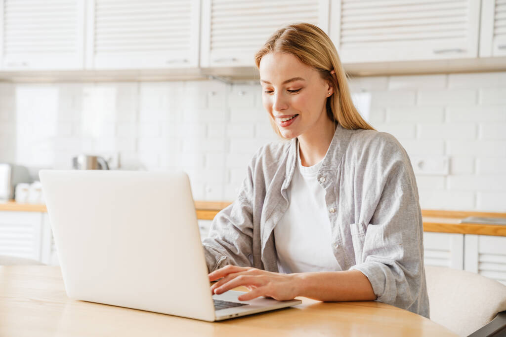 Vidám szőke lány mosolyog és használja laptop, miközben otthon ül konyha - Fotó, kép