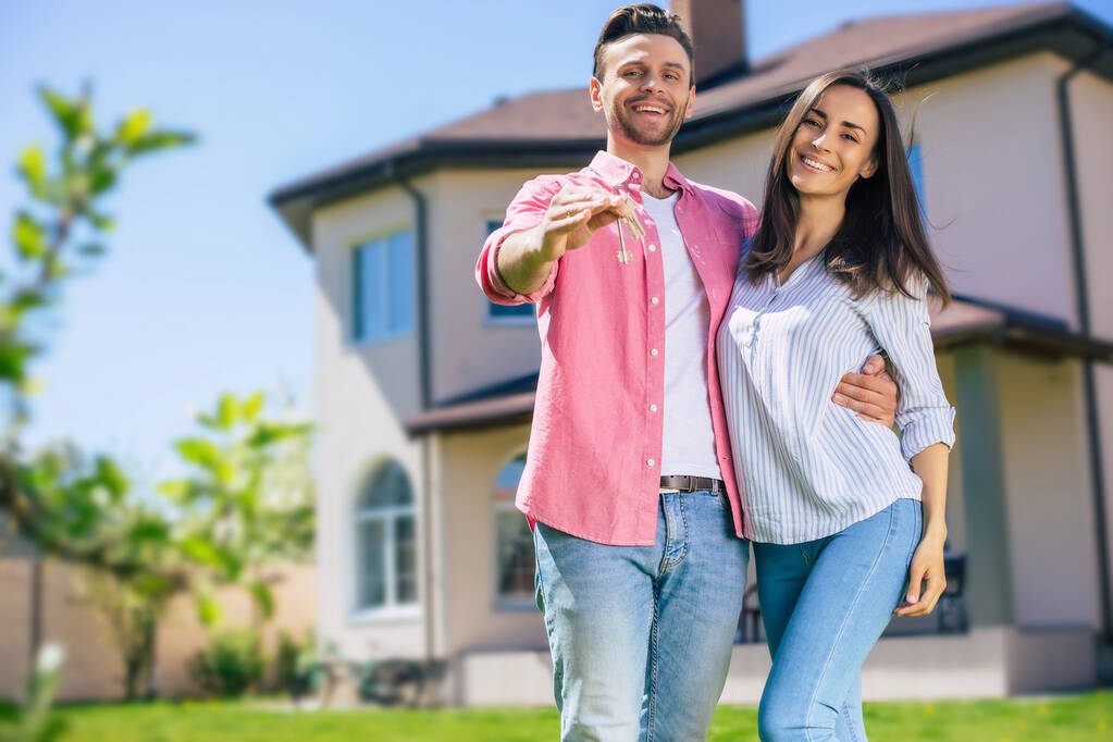 Modern beautiful excited young couple in love standing in front of the new big house with the keys in hands and hugging while they celebrating this buying - Photo, Image