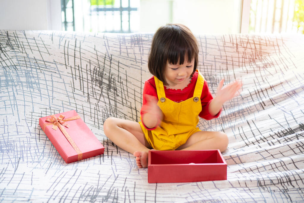 Dárek Překvapená mladá asijská dívka sedí na pohovce a otevírá dárek. Mockrát tleskám.. - Fotografie, Obrázek