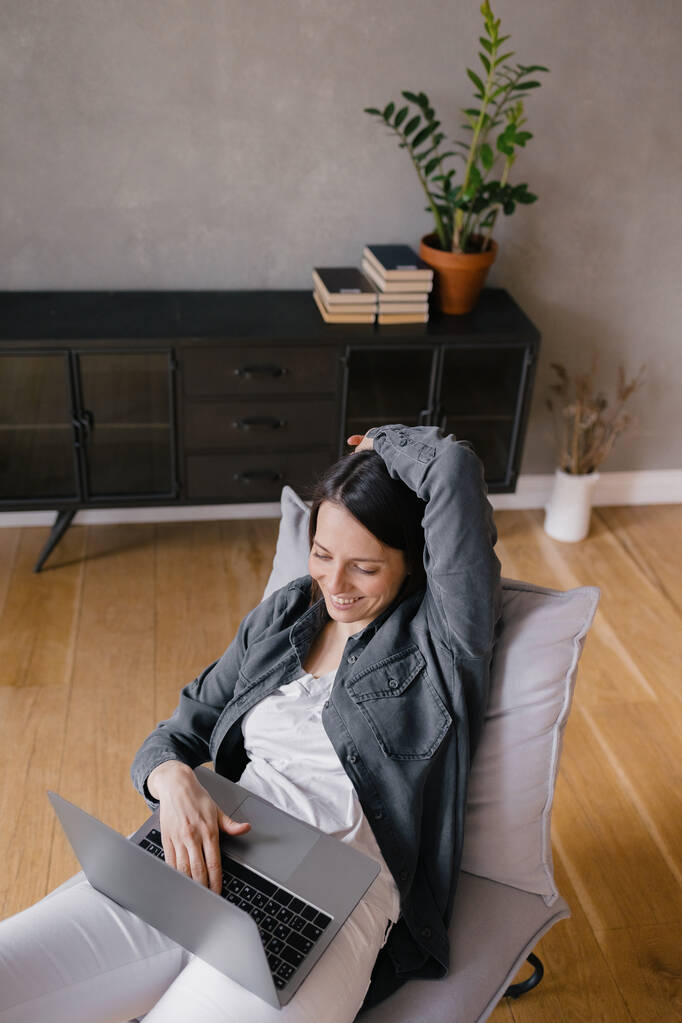 von oben, europäische Geschäftsfrau in grauem Hemd und weißer Jeans sitzt auf einem Stuhl, arbeitet mit einem Laptop und chattet am Telefon, SMS, surft im Internet oder erledigt Online-Einkäufe - Foto, Bild