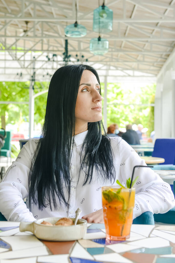Mooie jonge vrouw in het café met een glas cocktail en vanille-ijs. Brunette Kaukasisch meisje in restaurant alleen, sociaal afstandelijk concept als gevolg van pandemie. - Foto, afbeelding