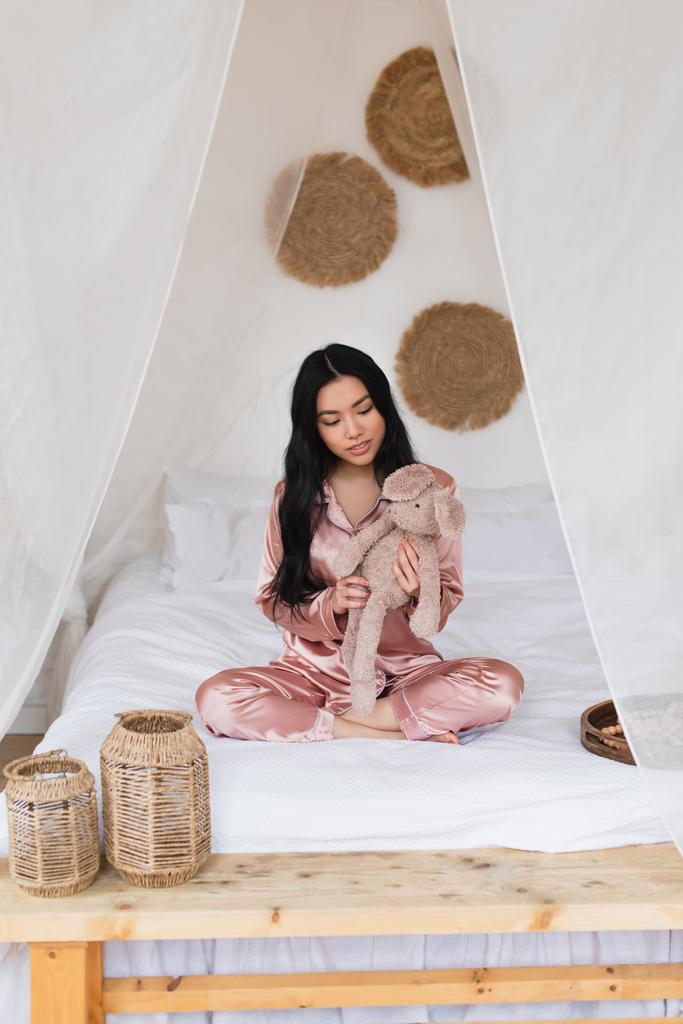 молода азіатка в шовковій піжамі сидить з схрещеними ногами і тримає м'яку іграшку в спальні
 - Фото, зображення