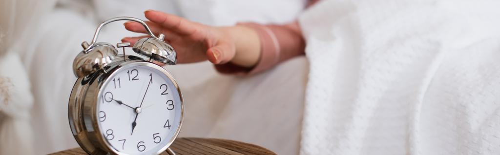 vista de cerca de la mano femenina extendiéndose a la mesita de noche con reloj despertador en el dormitorio, pancarta - Foto, Imagen