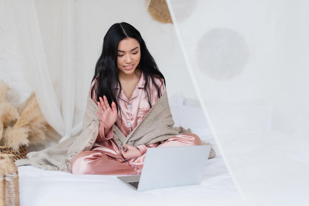 usmívající se mladá asijská žena sedí na posteli a mává rukou, zatímco s video hovor s notebookem v ložnici - Fotografie, Obrázek