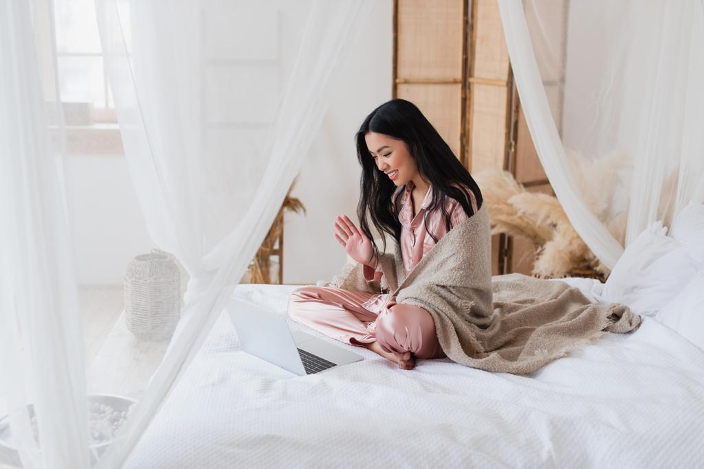 vrolijk jong aziatisch vrouw zitten op bed en zwaaien hand terwijl het hebben van video gesprek met laptop in slaapkamer - Foto, afbeelding