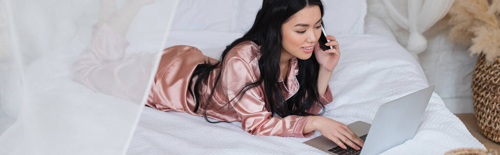 Nuori aasialainen nainen silkkipyjamassa makaa sängyssä, puhuu kännykkään ja katselee kannettavaa makuuhuoneessa, banneri - Valokuva, kuva