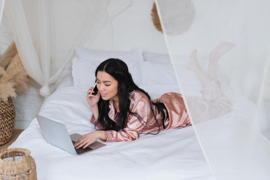 junge Asiatin im seidenen Pyjama liegt auf dem Bett, telefoniert und schaut auf Laptop im Schlafzimmer - Foto, Bild