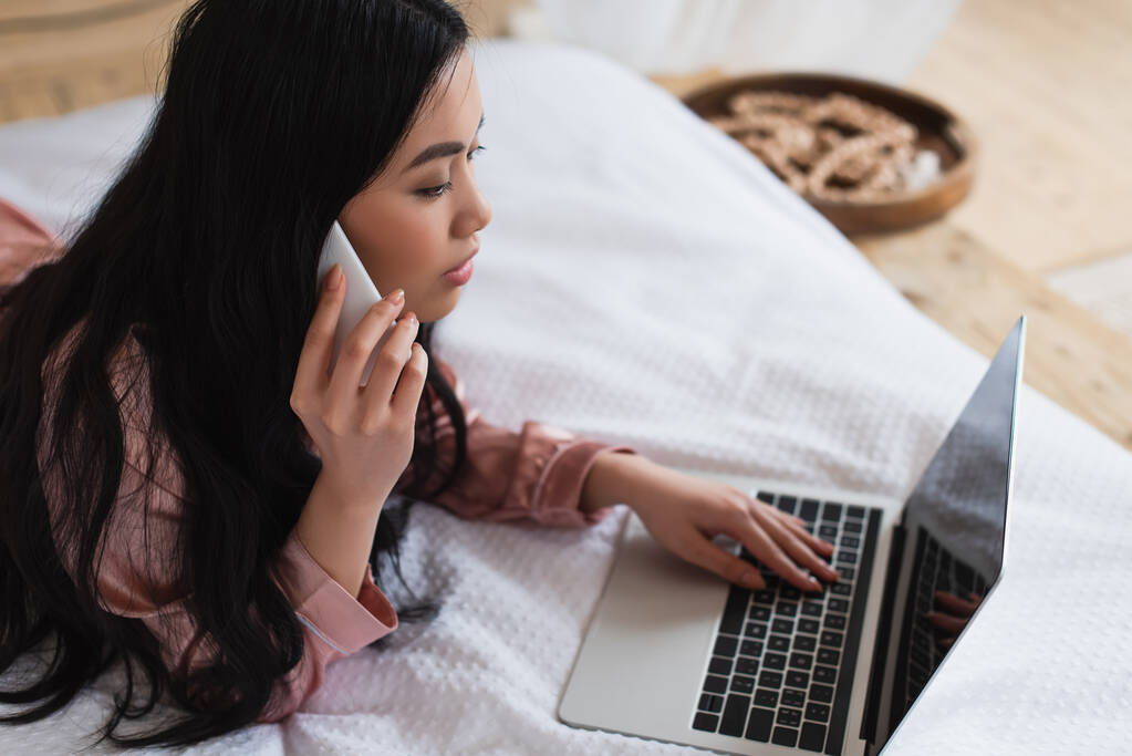 jovem mulher asiática em pijama de seda deitado na cama, falando no celular e olhando para laptop com tela em branco no quarto - Foto, Imagem