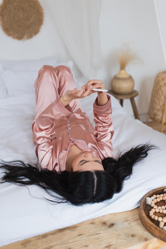 絹のパジャマを着た若いアジア人女性が寝室に携帯電話を持ってベッドに横になり - 写真・画像