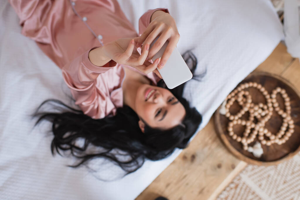 widok z góry uśmiechnięta młoda azjatycka kobieta w jedwabne piżamy leżące na łóżku i biorąc selfie z telefonu komórkowego w sypialni - Zdjęcie, obraz