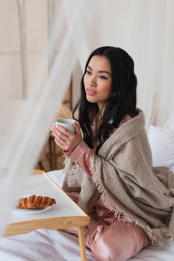 νεαρή Ασιάτισσα με μεταξωτές πιτζάμες και κουβέρτα που κάθεται στο κρεβάτι με ένα φλιτζάνι καφέ κοντά στο τραπέζι με κρουασάν στην κρεβατοκάμαρα - Φωτογραφία, εικόνα