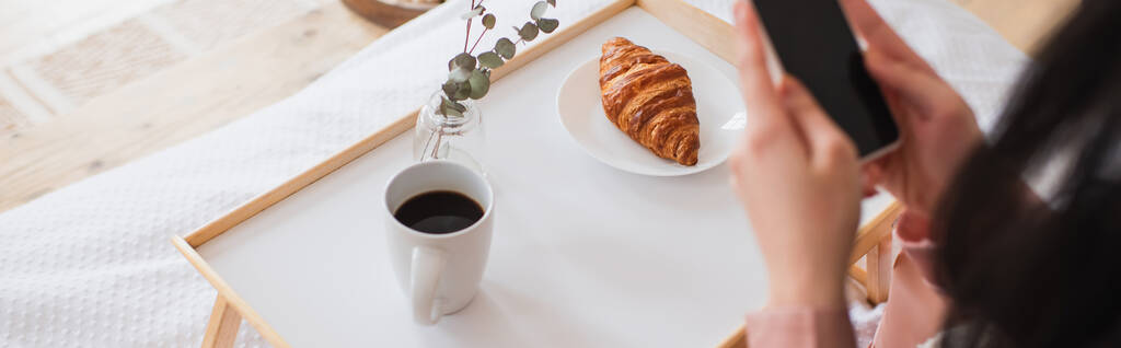 bijgesneden weergave van jonge brunette vrouw met mobiele telefoon in de buurt van tafel met koffie en croissant in de slaapkamer, banner - Foto, afbeelding