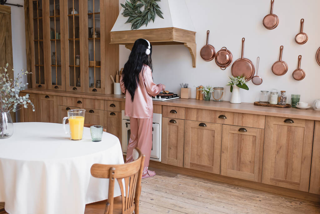 joven asiático mujer en rosa seda pijama y auriculares cocinar desayuno en cocina - Foto, Imagen