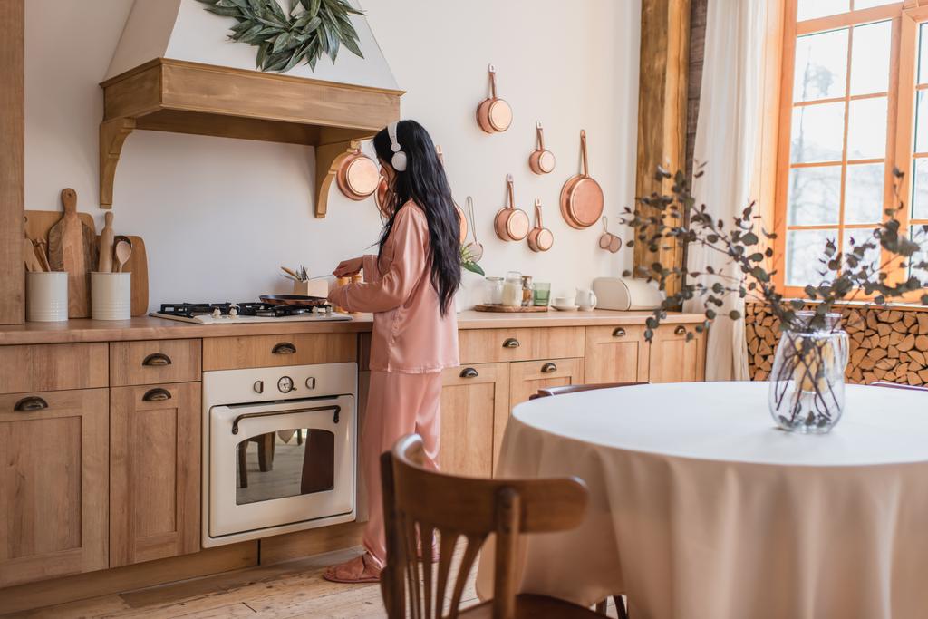 jonge aziatische vrouw in roze zijde pyjama en koptelefoon koken ontbijt in de buurt van tafel in de keuken - Foto, afbeelding