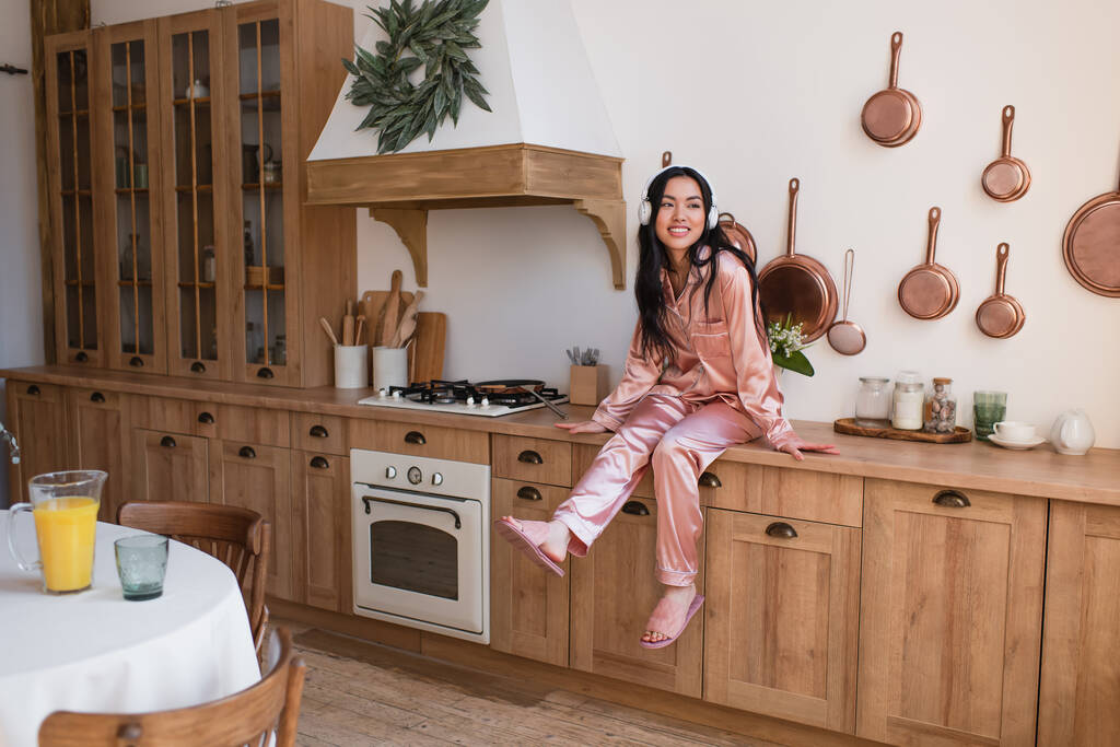 молодая азиатка в розовой шелковой пижаме и наушниках, сидящая рядом с плитой на кухне - Фото, изображение