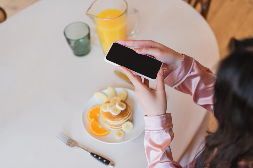 részleges kilátás fiatal nő gazdaság smartphone és fénykép a palacsinta gyümölcsök közelében narancslé a konyhában - Fotó, kép