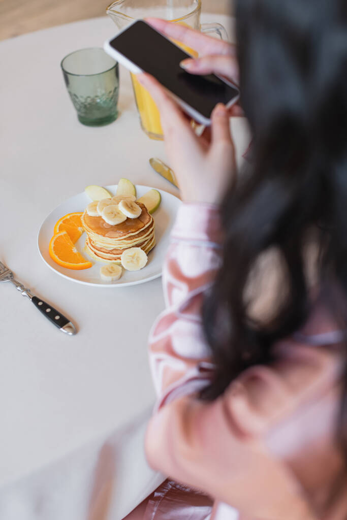abgeschnittene Ansicht einer jungen Frau, die ihr Handy in der Hand hält und Pfannkuchen mit Früchten und Orangensaft in der Küche fotografiert - Foto, Bild