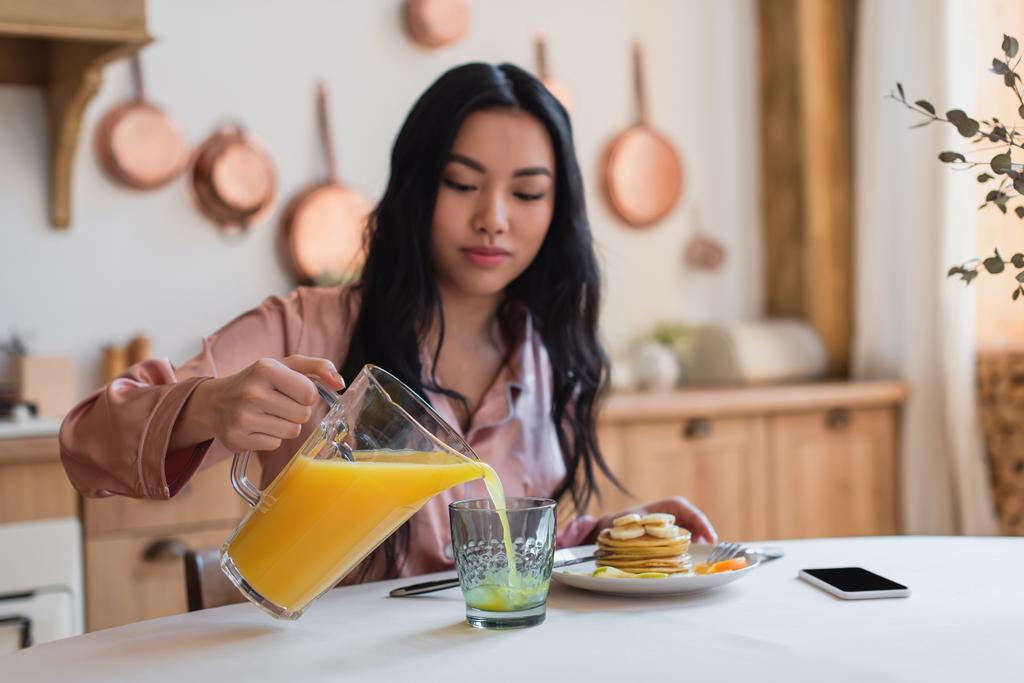 young asian woman in silk pajamas pouring orange juice into glass near pancakes and cellphone on table in kitchen - Photo, Image