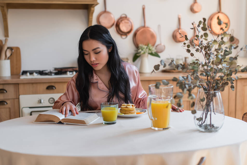 νεαρή Ασιάτισσα με μεταξωτές πιτζάμες διαβάζει το βιβλίο, ενώ τρώει πρωινό στην κουζίνα - Φωτογραφία, εικόνα