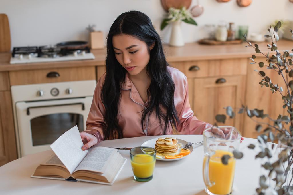 Nuori aasialainen tyttö silkki pyjama käsittelyssä kirja, kun ottaa herkullinen aamiainen keittiössä - Valokuva, kuva