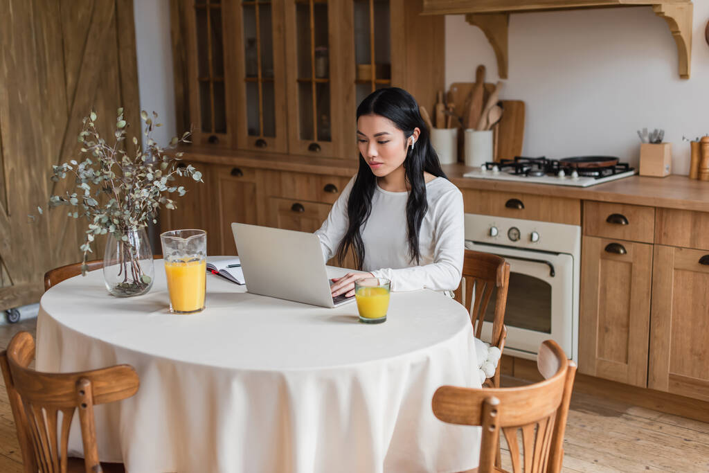 ernsthafte junge asiatische Frau mit Kopfhörern sitzt auf dem Tisch und benutzt Laptop in der Nähe von Notebook mit Stift in der Küche - Foto, Bild