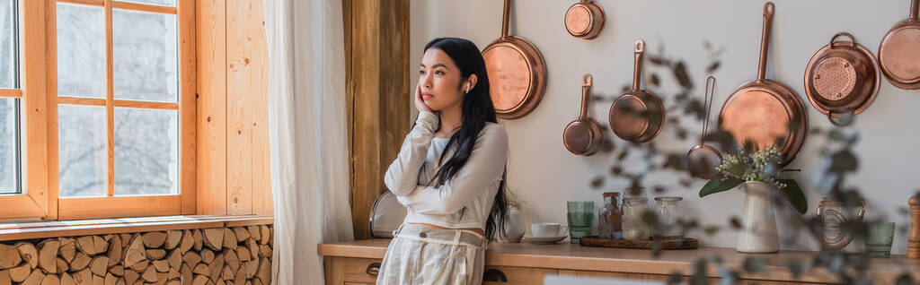 figyelmes fiatal ázsiai nő fülhallgató álló kézzel arccal a konyhában, banner - Fotó, kép