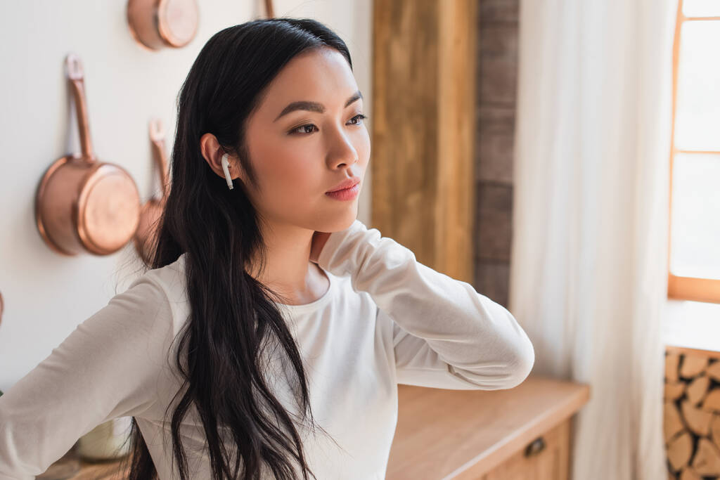 cansado joven asiático mujer en blanco blusa de pie con auriculares y celebración de la mano cerca del cuello en la cocina - Foto, imagen