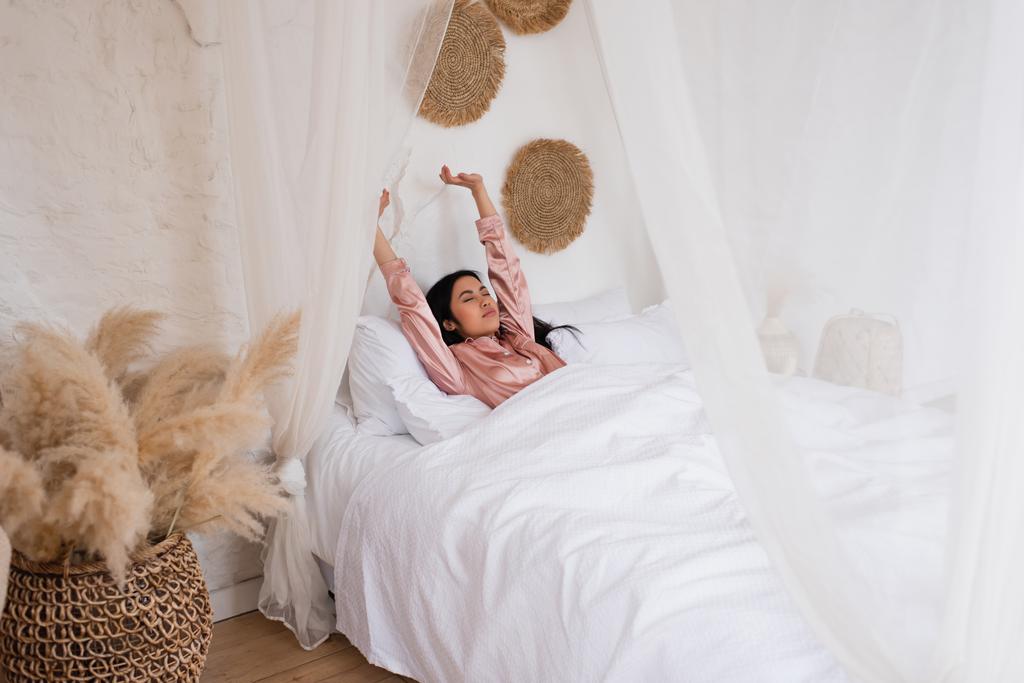 сонна молода азіатська жінка в шовковій піжамі прокидається з простягнутими руками в спальні
 - Фото, зображення