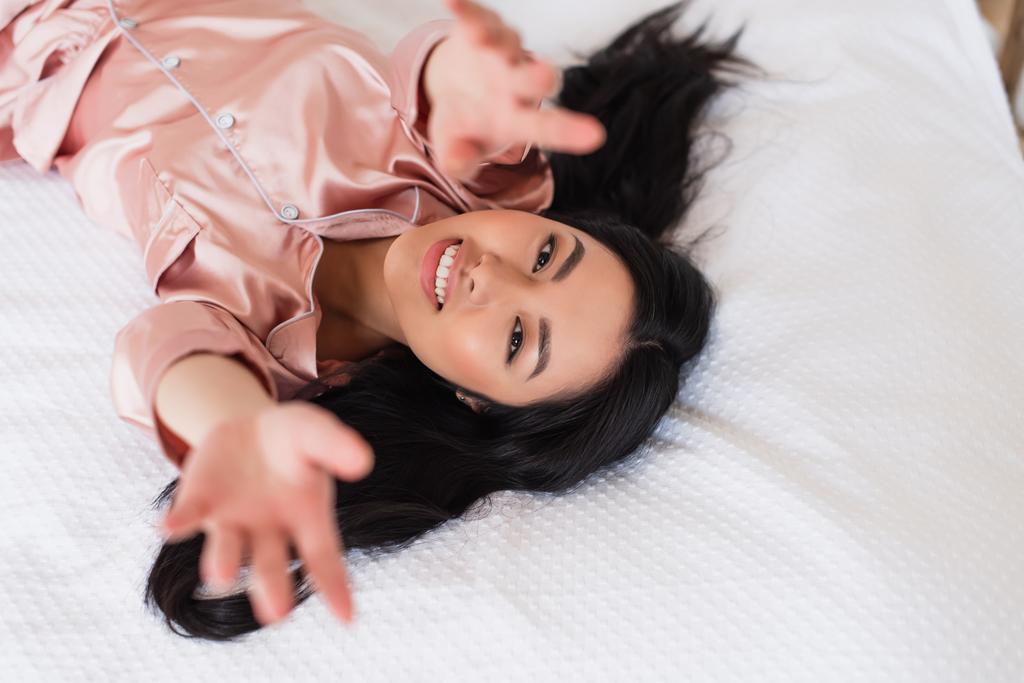 bovenaanzicht van jonge aziatische vrouw liggend op wit linnen met uitgestrekte handen en kijkend naar camera in slaapkamer - Foto, afbeelding